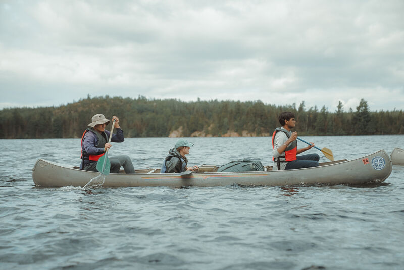 The image shows three people in a canoe on a lake. The person in the front is wearing a hat and paddling. The person in the middle is also wearing a hat. The person in the back is paddling as well. All three are wearing life vests. The canoe is in the middle of the lake, and there are trees in the background. The sky is cloudy.